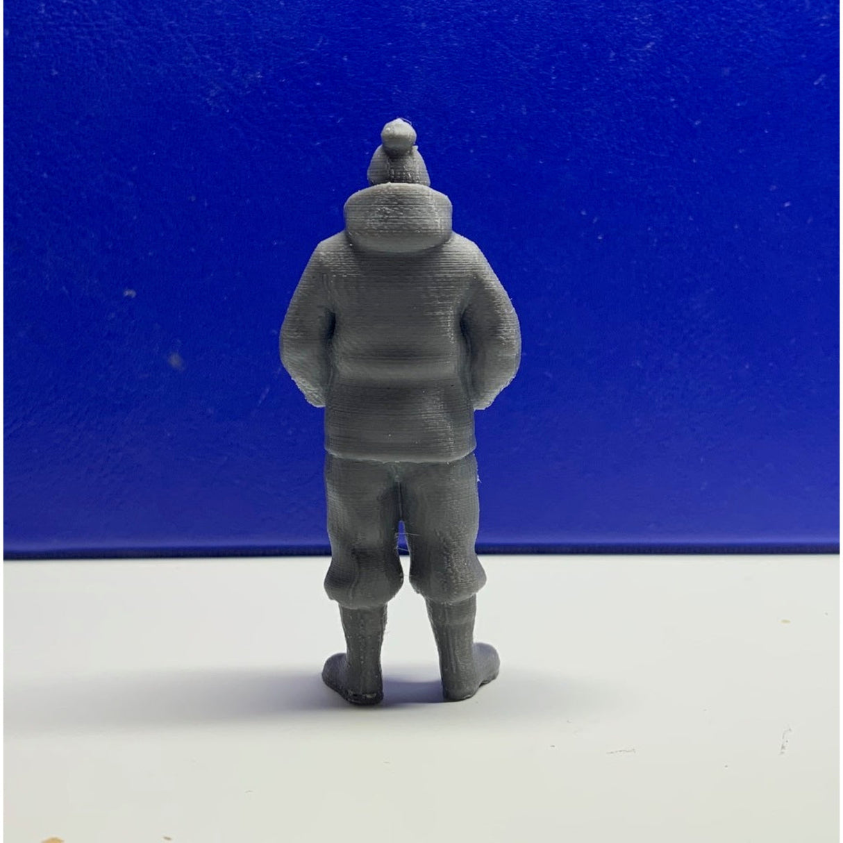 Rear view of unpainted grey 3D printed trawler fisherman figure wearing waterproof jacket, boots, and bobble hat. Model Boat Crew figure